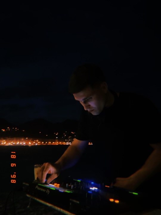Me at a DJ controller at night, a town's lights along the water behind me.