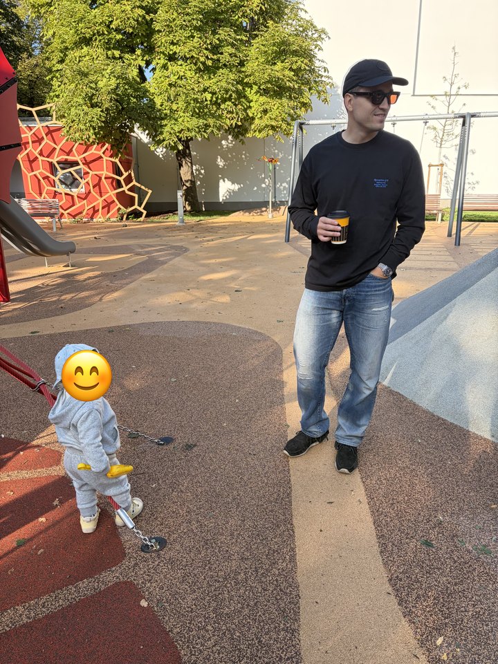 Me with a coffee at a playground, my son in a grey tracksuit and cap beside me, his face covered with an emoji.